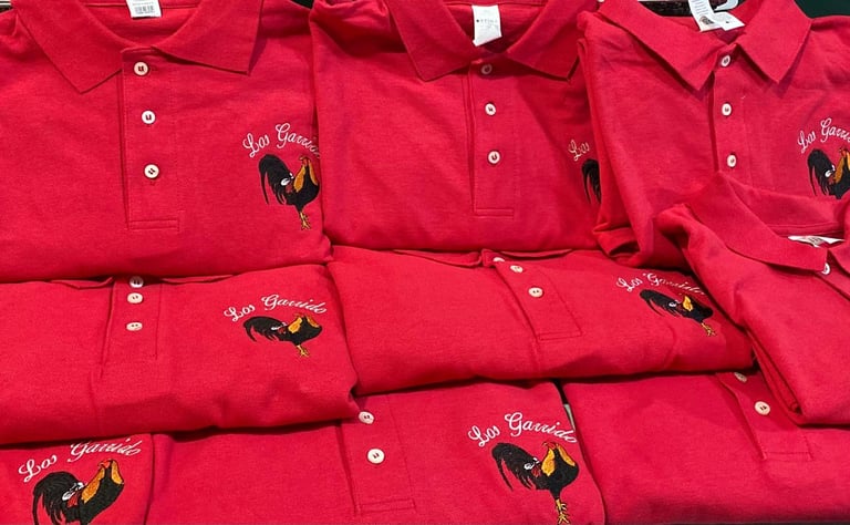 Múltiples playeras polo rojas brillantes dobladas y apiladas con logo de gallo y texto bordado