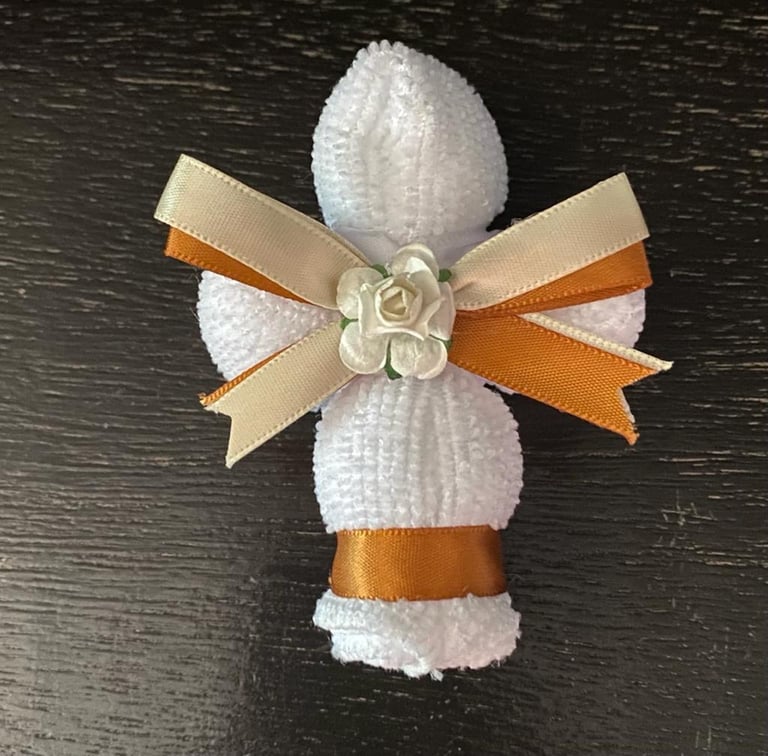Handmade cross decoration made from white textured material with cream and orange ribbon bow and small white flower center on dark wood surface