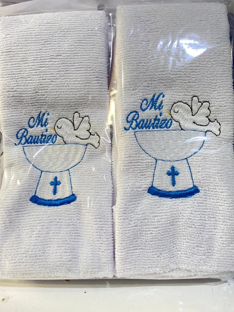 Two packaged white towels with embroidered Mi Bautizo designs featuring a smiling baby character and blue cross motif